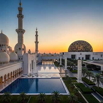 A grand mosque courtyard at sunset with reflective pools, white marble domes, tall minarets, and a modern patterned dome in the background.