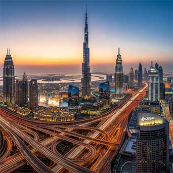Dubai skyline at sunset with the Burj Khalifa towering above illuminated skyscrapers and a network of glowing highways below.